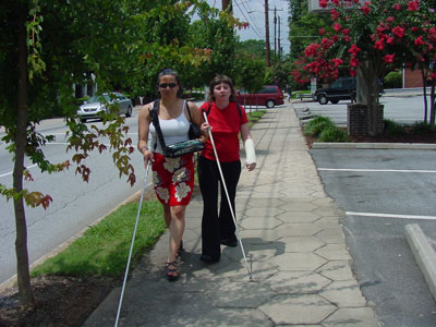 Two women, both with canes, are walking down a sidewalk. One woman is carrying a BrailleNote.