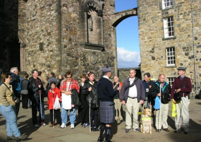 A guide in a blue-and-gray kilt speaks to a group of people, many with canes and one with a dog guide, in front of castle walls.