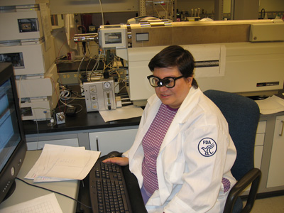 Photo of Judy Summers-Gates sitting at a computer displaying various graphs. She is wearing eyeglasses with a bioptic telescope.