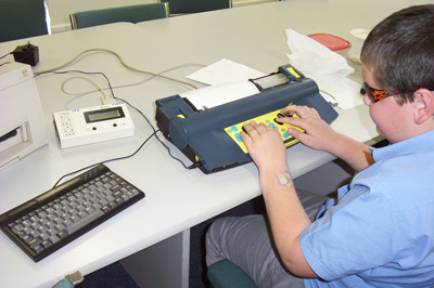 Photo of a boy using a Mountbatten Brailler that is connected to a QWERTY keyboard, a MIMic display, and a printer.