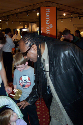 Photo of Stevie Wonder talking to some young children in front of a HumanWare sign.