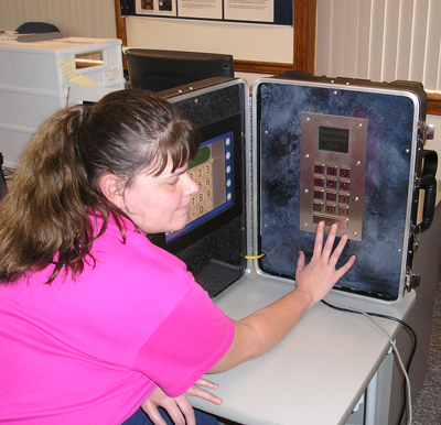 Photo of a woman pressing the accessibility button of a keypad mounted on the inside of a suitcase. The touch screen is mounted on the other side of the suitcase.