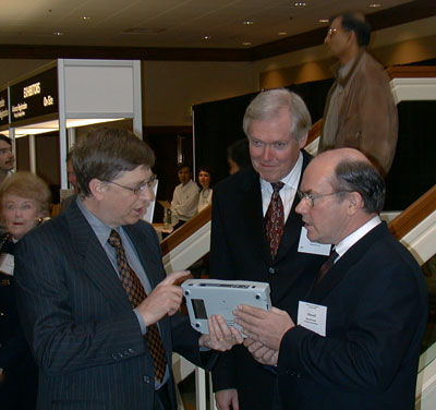 Russell Smith holds a BrailleNote as an interested Bill Gates points to it and speaks. Jim Halliday smiles.