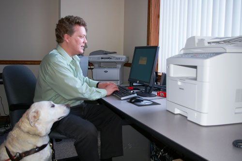 Photo of a man typing on a computer next to the Samsung desktop unit, while a dog guide observes.