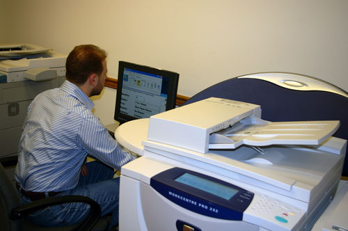 Photo of a man working at a computer screen next to a Xerox WorkCentre Pro 232 photocopier. The menu on the computer screen offers choices for quantity, duplex, tray, staples, and collation.