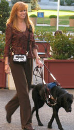 Photo of a woman walking outside with a dog guide. She is wearing the BrailleNote PK slung across her shoulder on a strap.