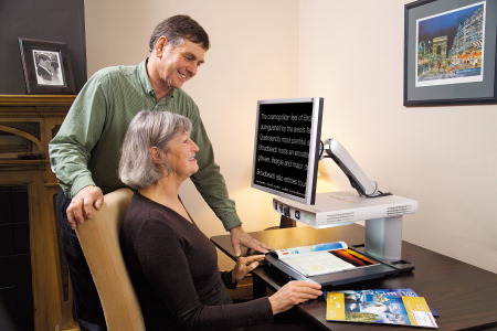 A seated woman reads magnified text in high contrast. A man, standing behind her, watches over her shoulder.