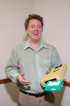 A photo of Darren holding the huge old NLS cassette player and a box of cassettes in one arm, with the tiny iPod shuffle in his ear.