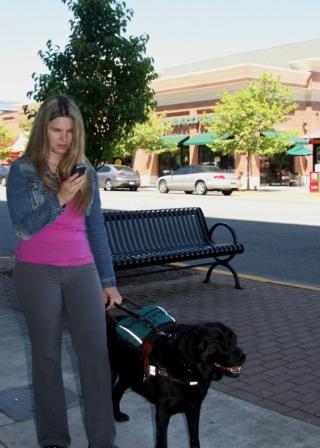 A woman holding the dog guide's harness in her left hand and the Smartphone in her right hand. There is a Starbucks in the background.
