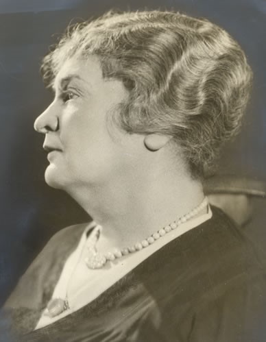 This head and shoulders photographic portrait of Anne Sullivan Macy shows her in profile, circa 1920. Anne's thick, glossy hair is short and waved. She is wearing a dark, embroidered V-neck garment with a light-colored blouse or collar underneath. The collar appears to be fastened with a decorative pin. She has a strand of pearls around her neck.