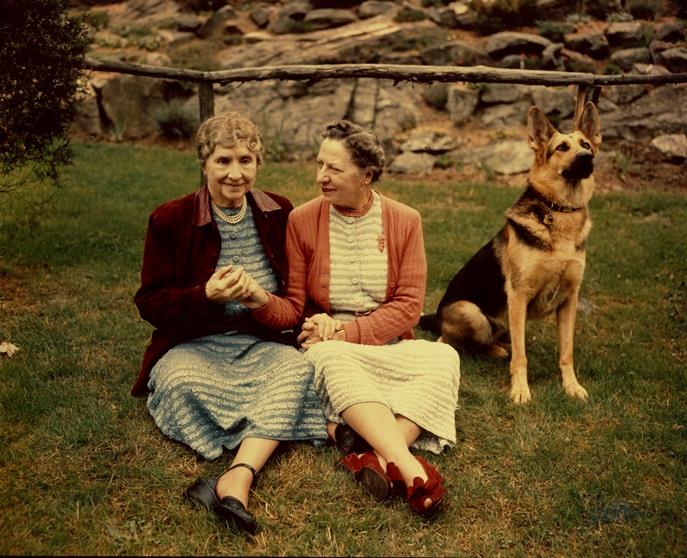 Helen Keller and Polly Thomson, seated outside on the grass with German Shepherd dog nearby, circa 1955