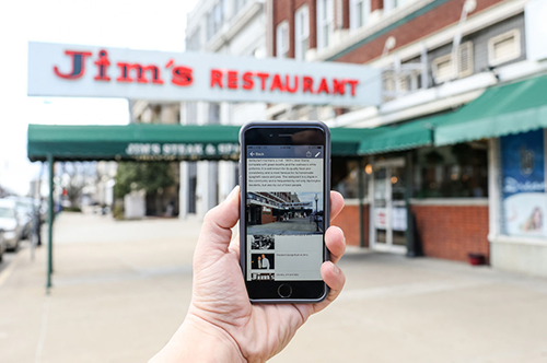 In the foreground, hand holding smartphone. Phone screen shows a landmark navigation app. In background is the landmark location, a restaurant with awning. Text on awning reads, "Jim's Restaurant"