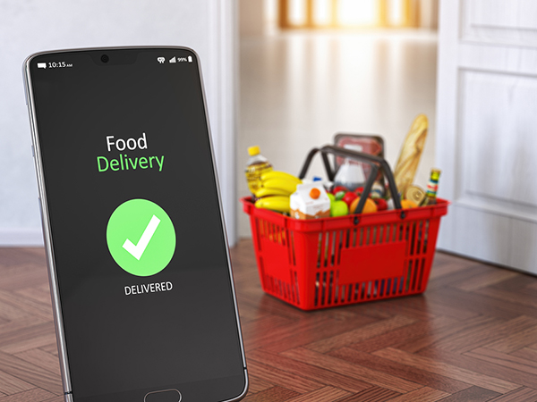 Smartphone in the foreground with a generic food delivery app on the screen. In the background is a shopping basket of food.