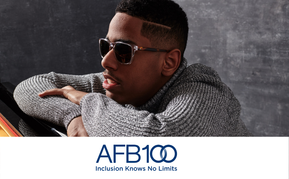 AFB 100. Inclusion knows no limits. Matthew Whitaker, a young black man wearing dark glasses and a grey turtleneck sweater sits in front of and is leaning onto a piano.