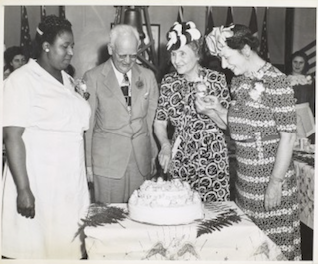 Mellissa Vail, Peter J. Salmon, Helen Keller and Polly Thomson are standing together. Keller is about to cut a decorated two-tiered cake that is on the table infront of them. 
