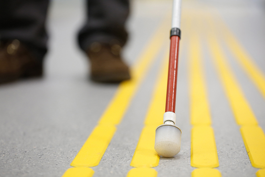 A person using a white cane walks on pavement with tactile markers.