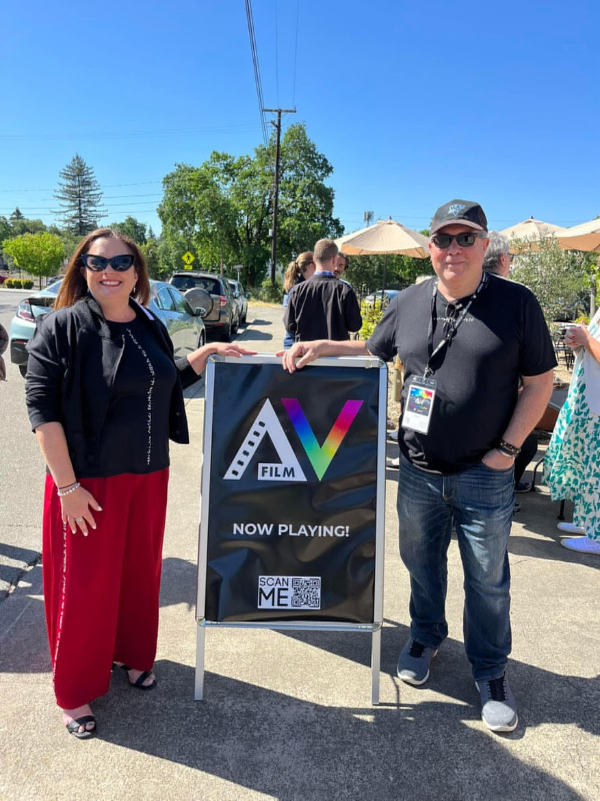From left to right: Melody Goodspeed and Bill Sarine. They stand next to a sign that reads AV Film, Now Playing!