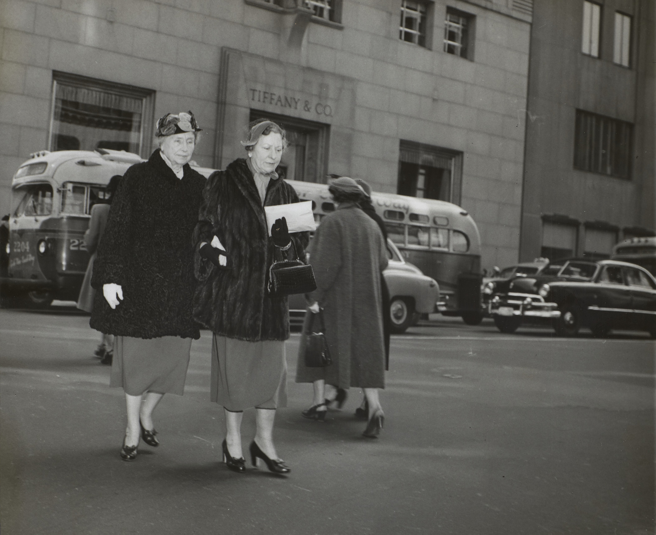 Helen Keller and Polly Thomson dressed in classy coats and heels, crossing a street in New York City. Pictured behind them are vintage cars and buses, and a tall NYC building.