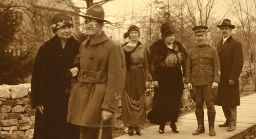 photograph from 1919, just after World War I. Helen Keller has the arm of a newly blinded soldier. He has a bandage over his left eye and is using a cane to help him walk. They are walking down a woodsy path in Baltimore, Maryland. Behind them is a stone wall and a large house with many windows, which is the Red Cross Institute for the Blind. Bringing up the rear, also on the path, are Annie Sullivan, Polly Thomson, and two other men, one in a military uniform.